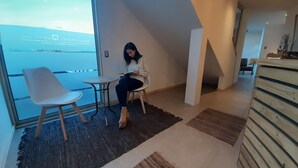 A modern interior with a woman sitting on a chair by a large window, a small table, and a wooden cabinet. - Apart Hotel Quillango (Punta Arenas)
