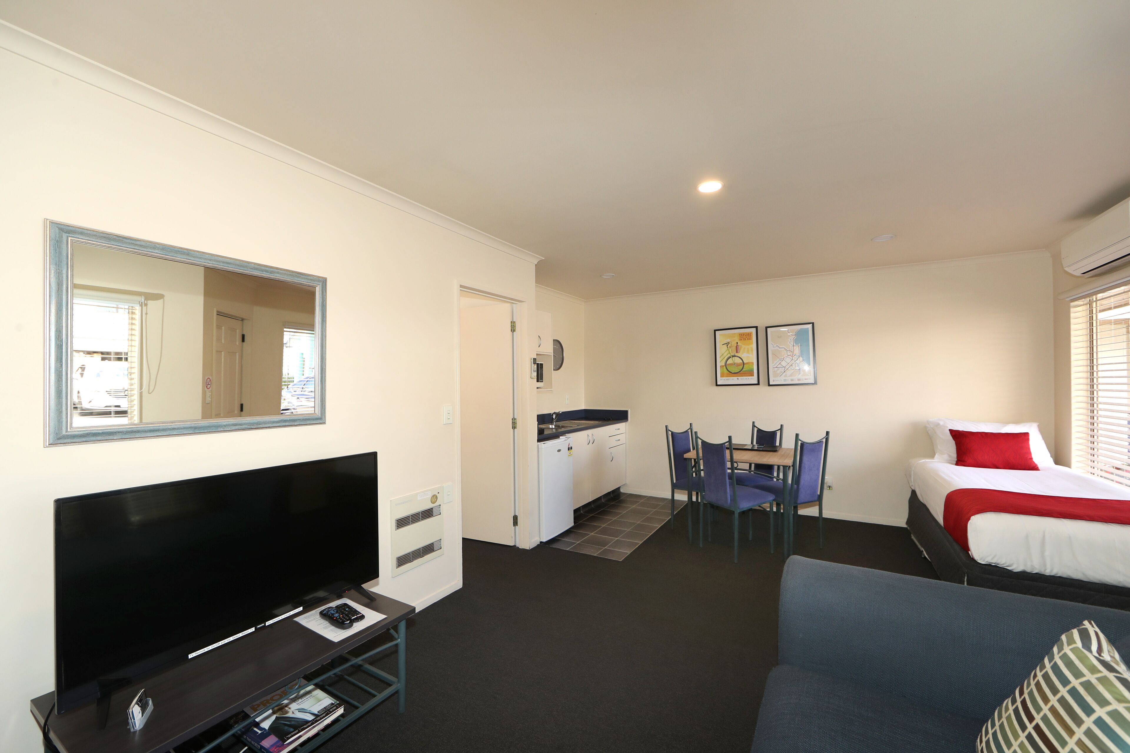 A hotel room with a large flat-screen TV, a dining area with chairs and a table, a bed with red bedding, and a small kitchenette.