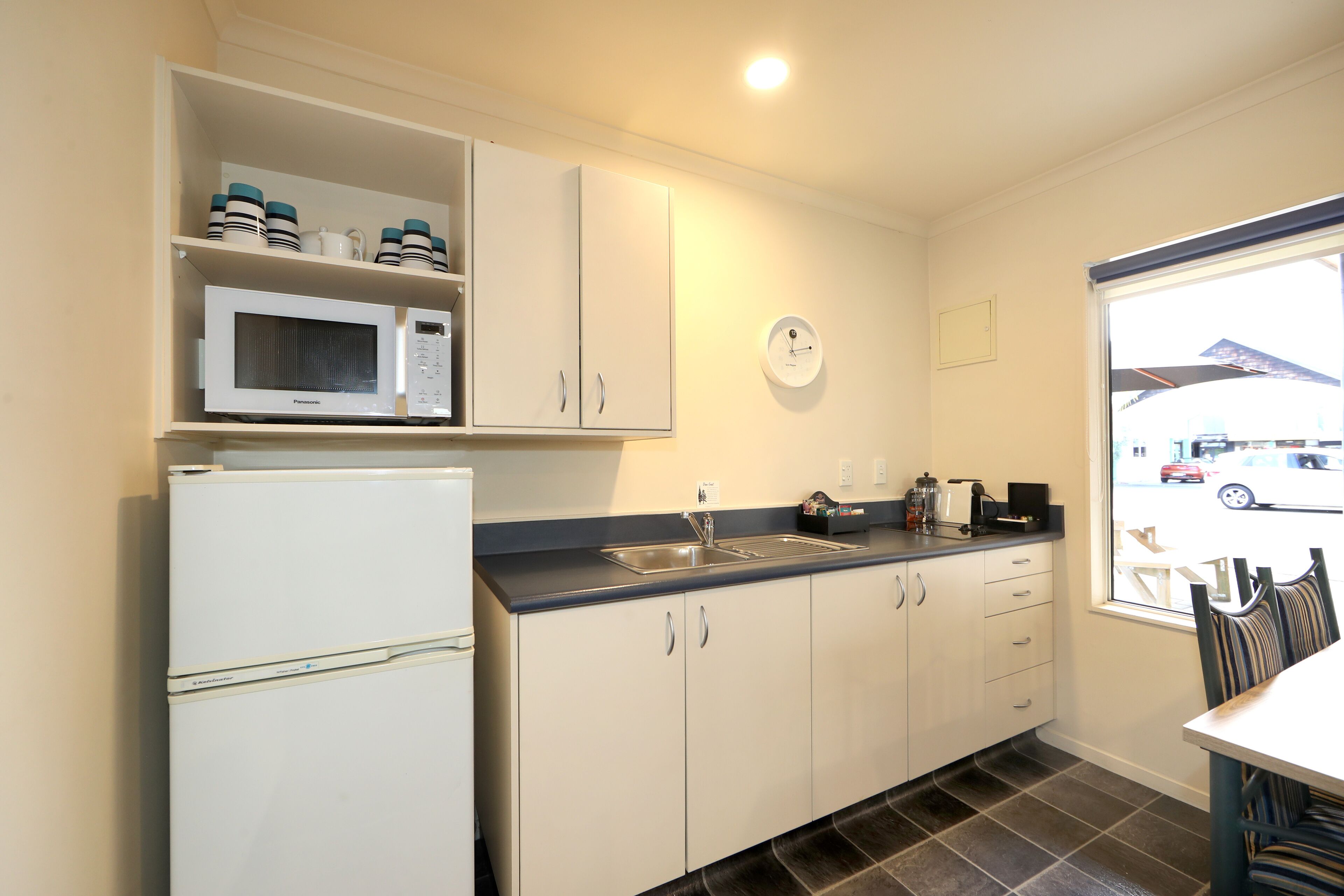 A compact kitchen with a refrigerator, microwave, and sink. There is a clock on the wall and a window with a view of the street.