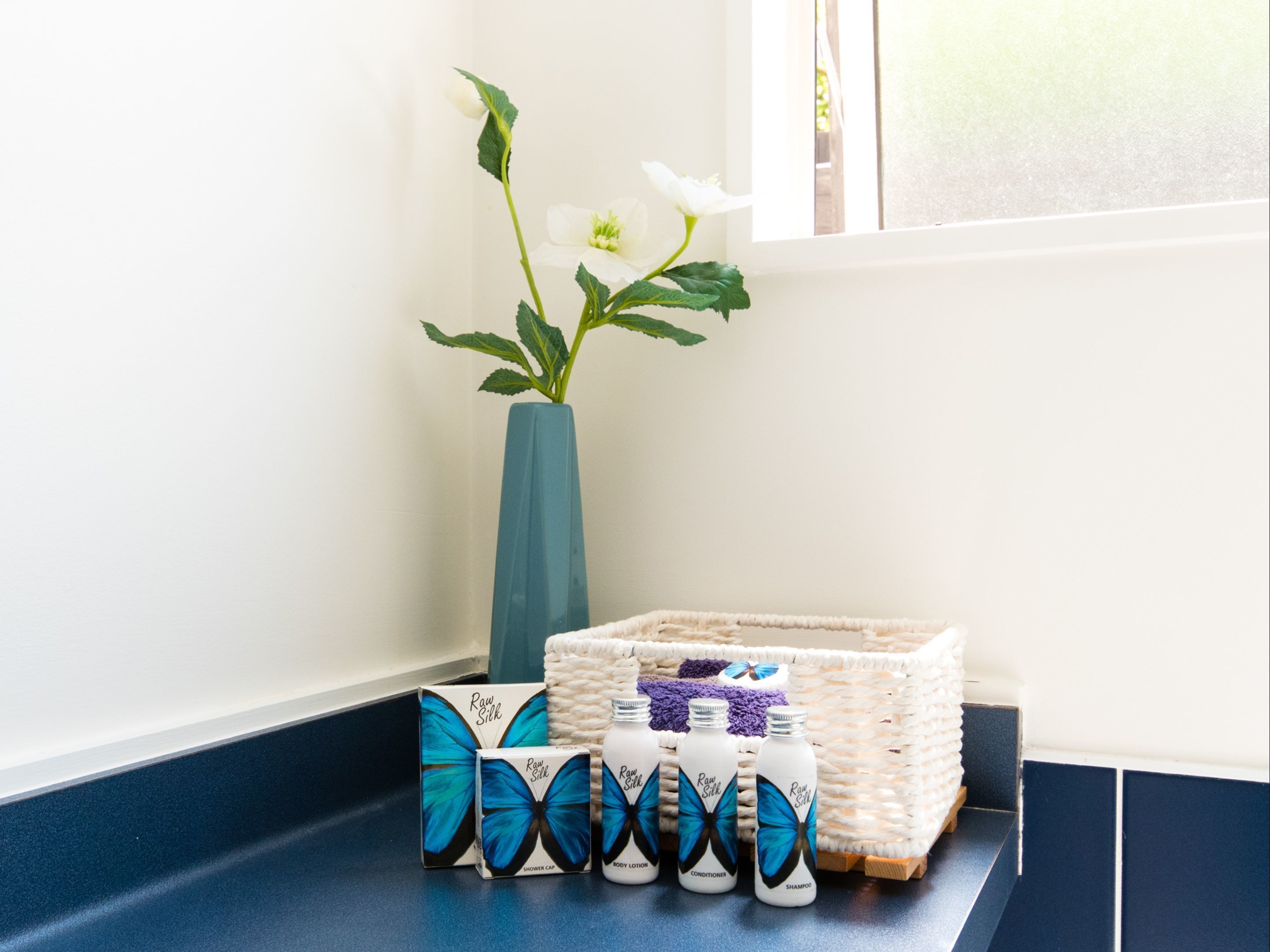 A bathroom with a blue vase, a woven basket, and three bottles of 'Raw Silk' products.