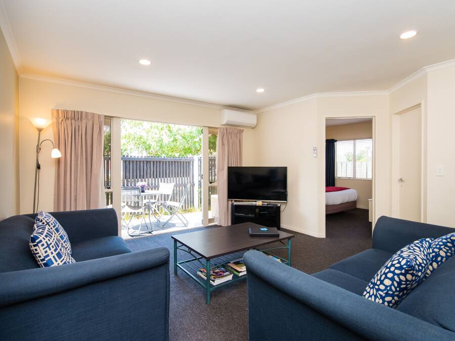 A living room with a sofa, a coffee table, and a TV. There is a sliding glass door leading to an outdoor area with a table and chairs.