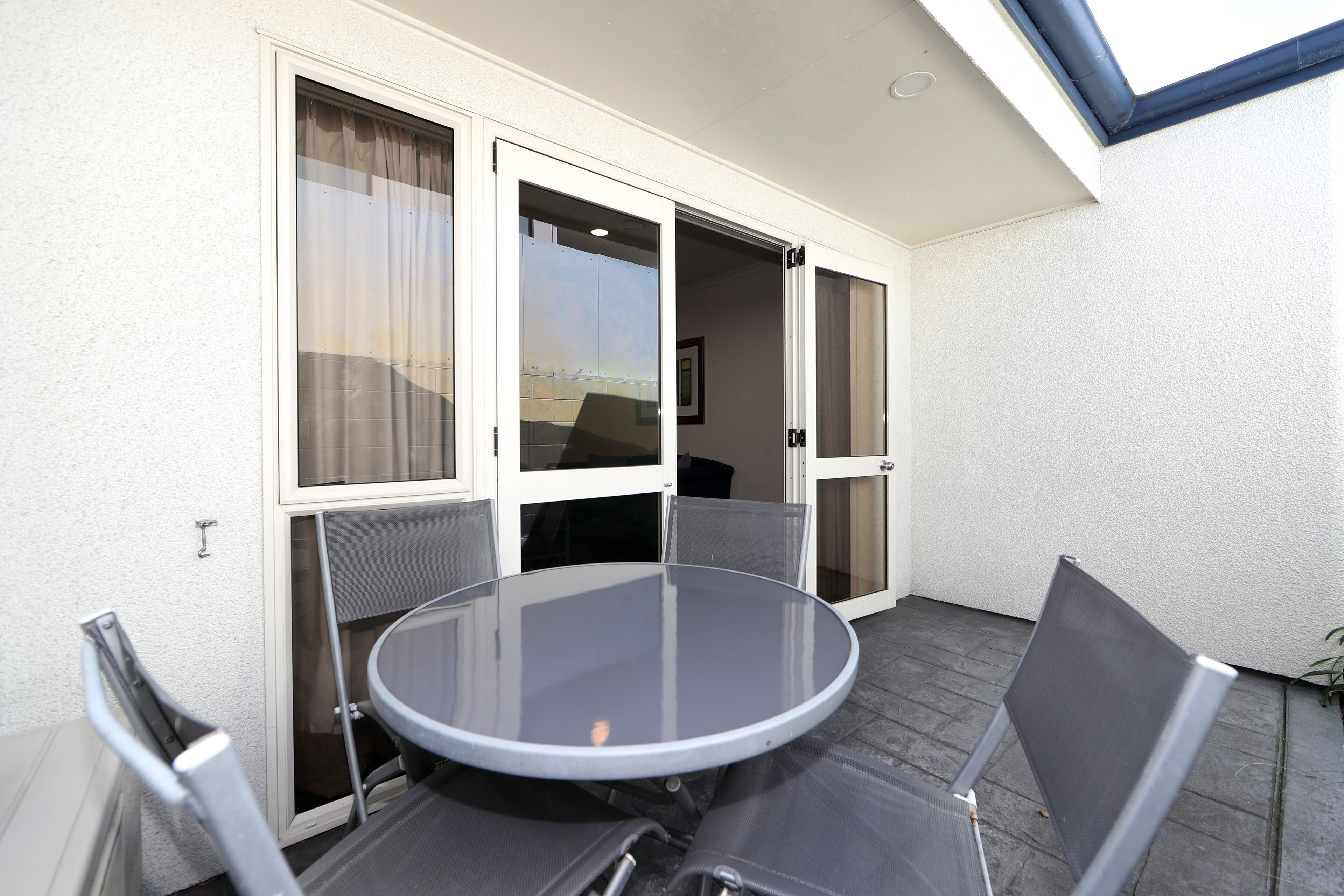 A small, enclosed balcony with a round glass table and two chairs, a sliding glass door, and a view of the outside.