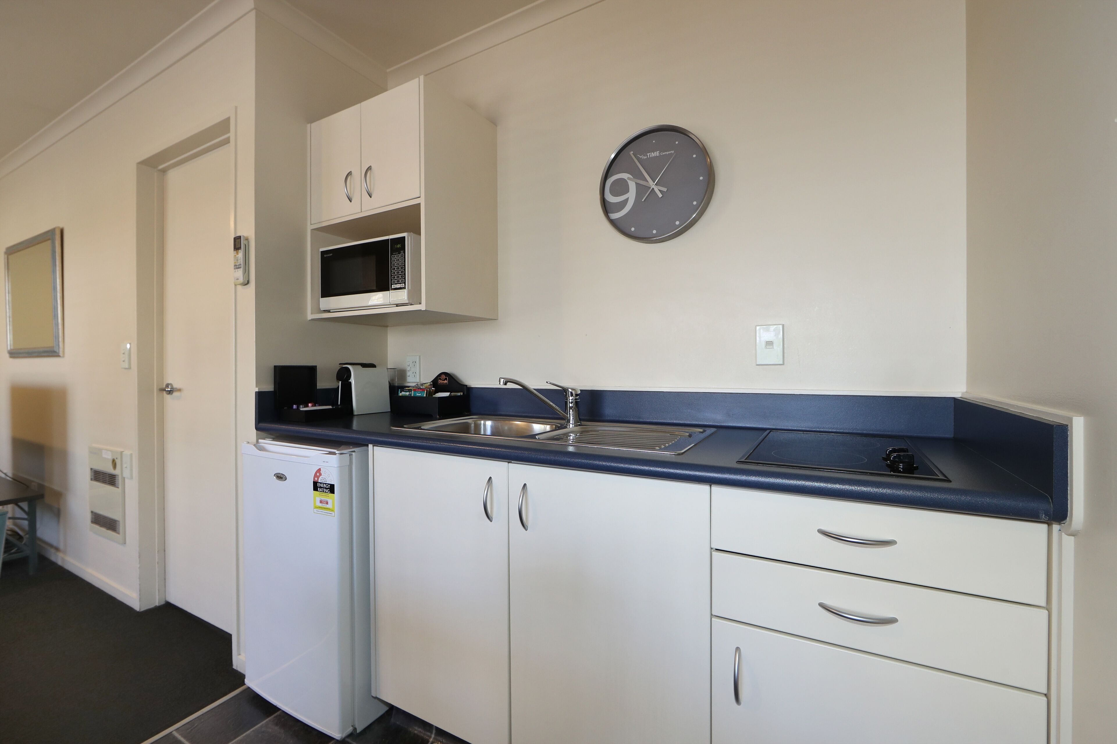 A compact kitchen with white cabinets, a stainless steel sink, a microwave, and a clock on the wall.
