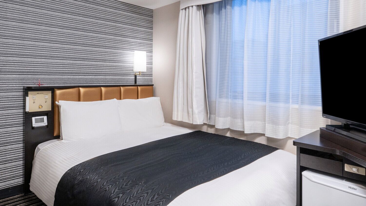 A hotel room with a bed, a television, a desk, and a wall-mounted control panel.