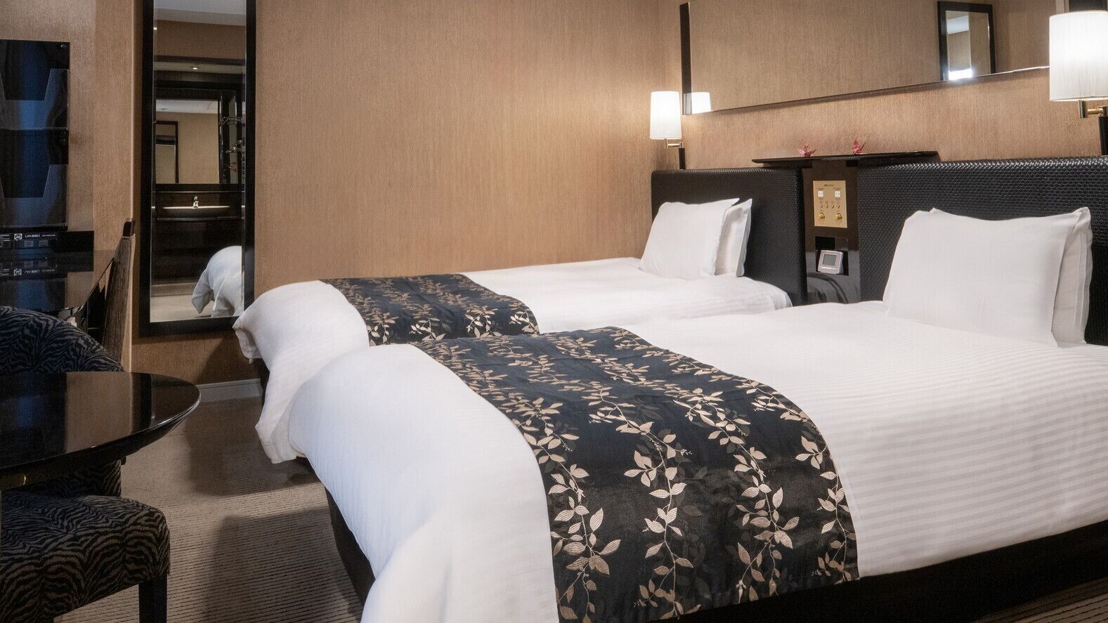 A hotel room with two beds, a desk, a chair, a mirror, and a wall-mounted control panel.