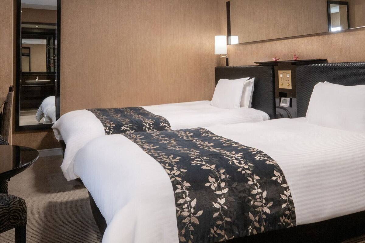 Ein Hotelzimmer mit zwei Betten, einem Schreibtisch, einem Sessel, einem Spiegel und einer wandmontierten Steuerung.