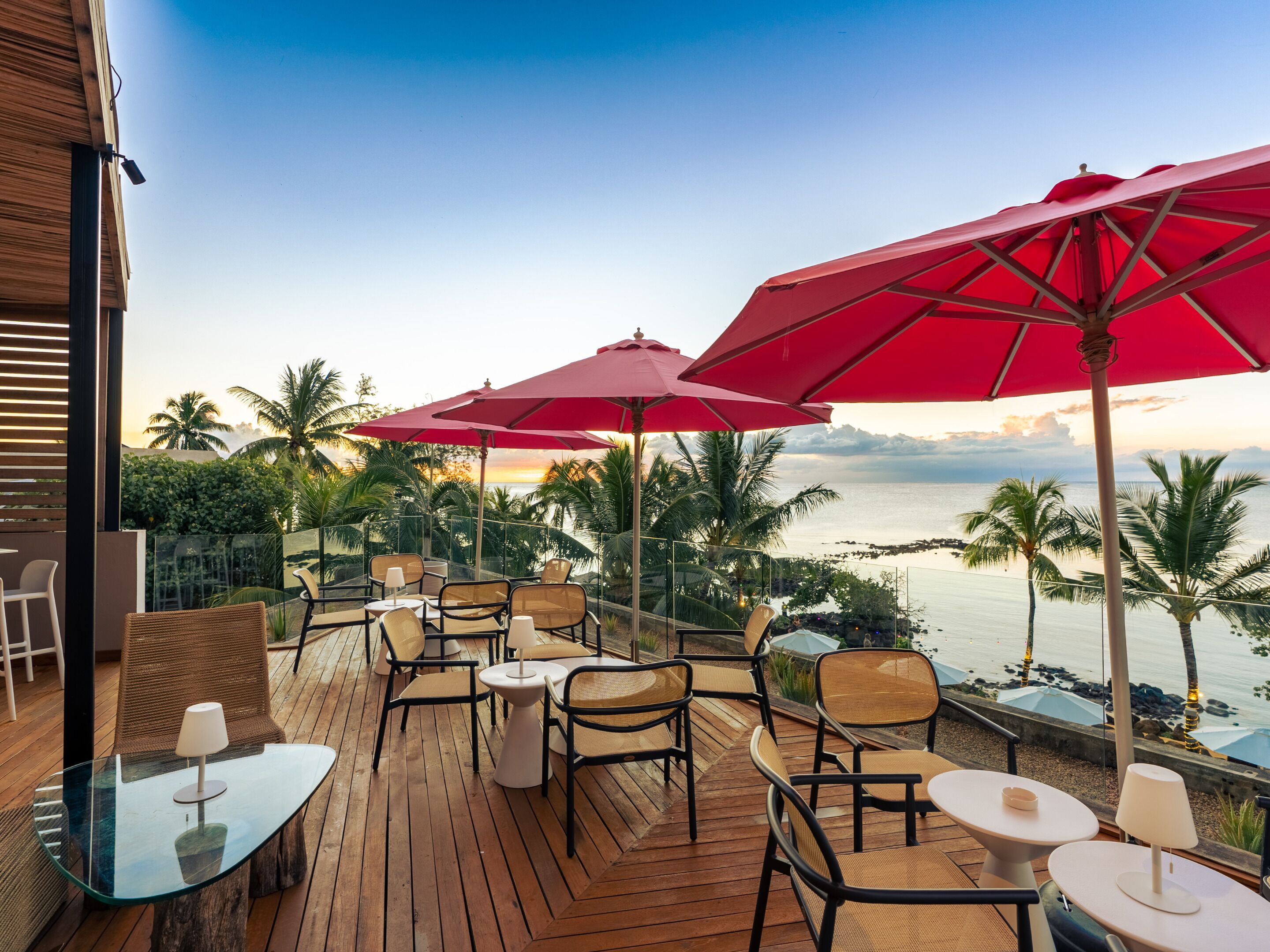 A wooden deck with outdoor seating, red umbrellas, and a view of the ocean.
