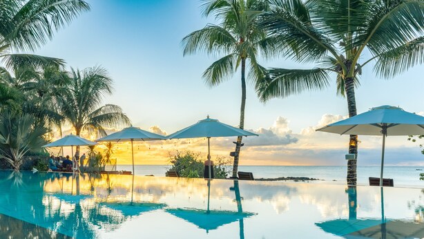 Zona de piscina con sombrillas, palmeras y vista al océano al atardecer.