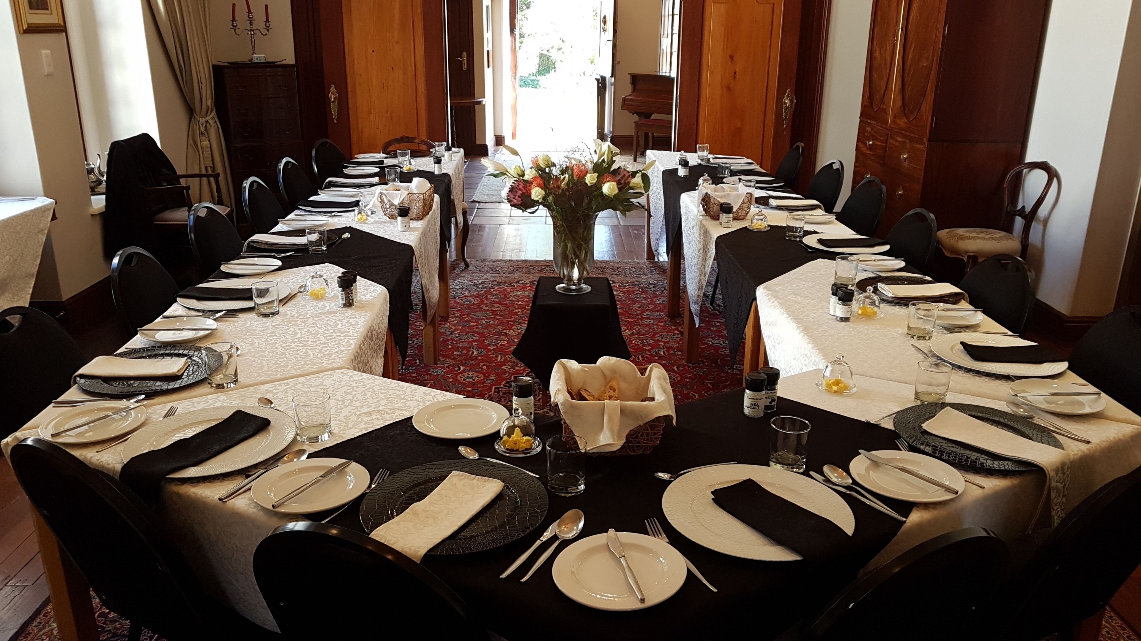 Una larga mesa de comedor preparada para una comida formal, con múltiples sillas, un jarrón de flores y un piano al fondo.