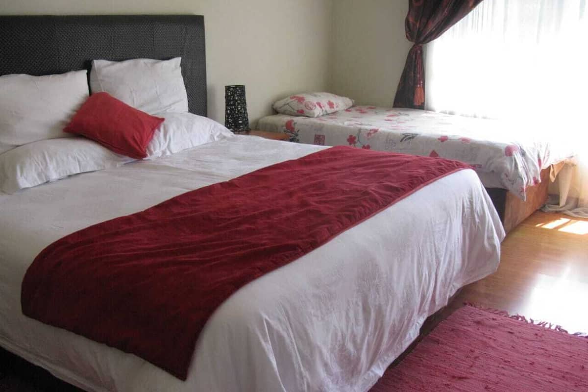 Una cama doble con una colcha roja y ropa de cama blanca, una cama más pequeña con ropa de cama floral, y una mesita de noche con una lámpara.