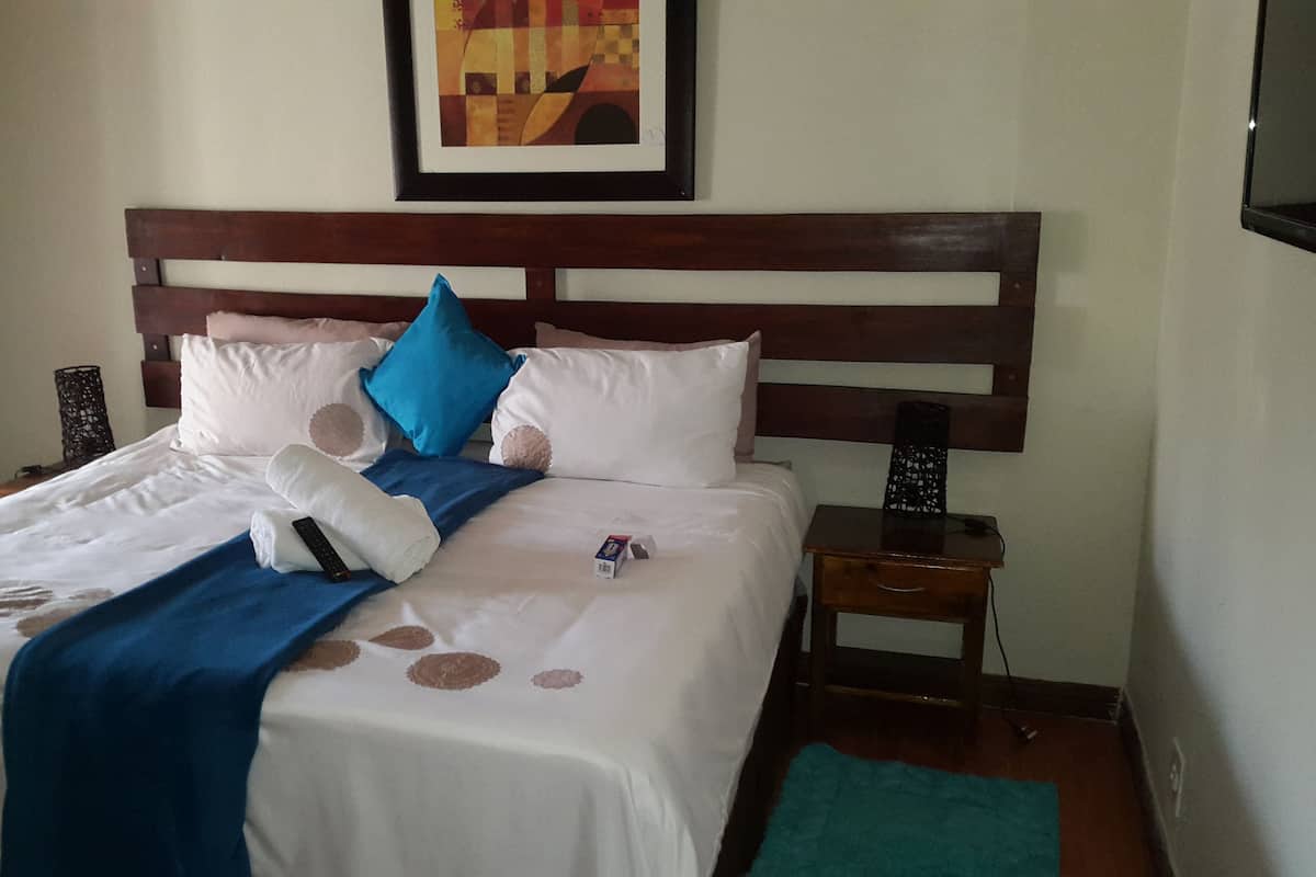 Una cama bien hecha con una colcha azul, almohadas blancas y un cabecero de madera. Hay una mesita de noche pequeña con una lámpara y un enchufe.