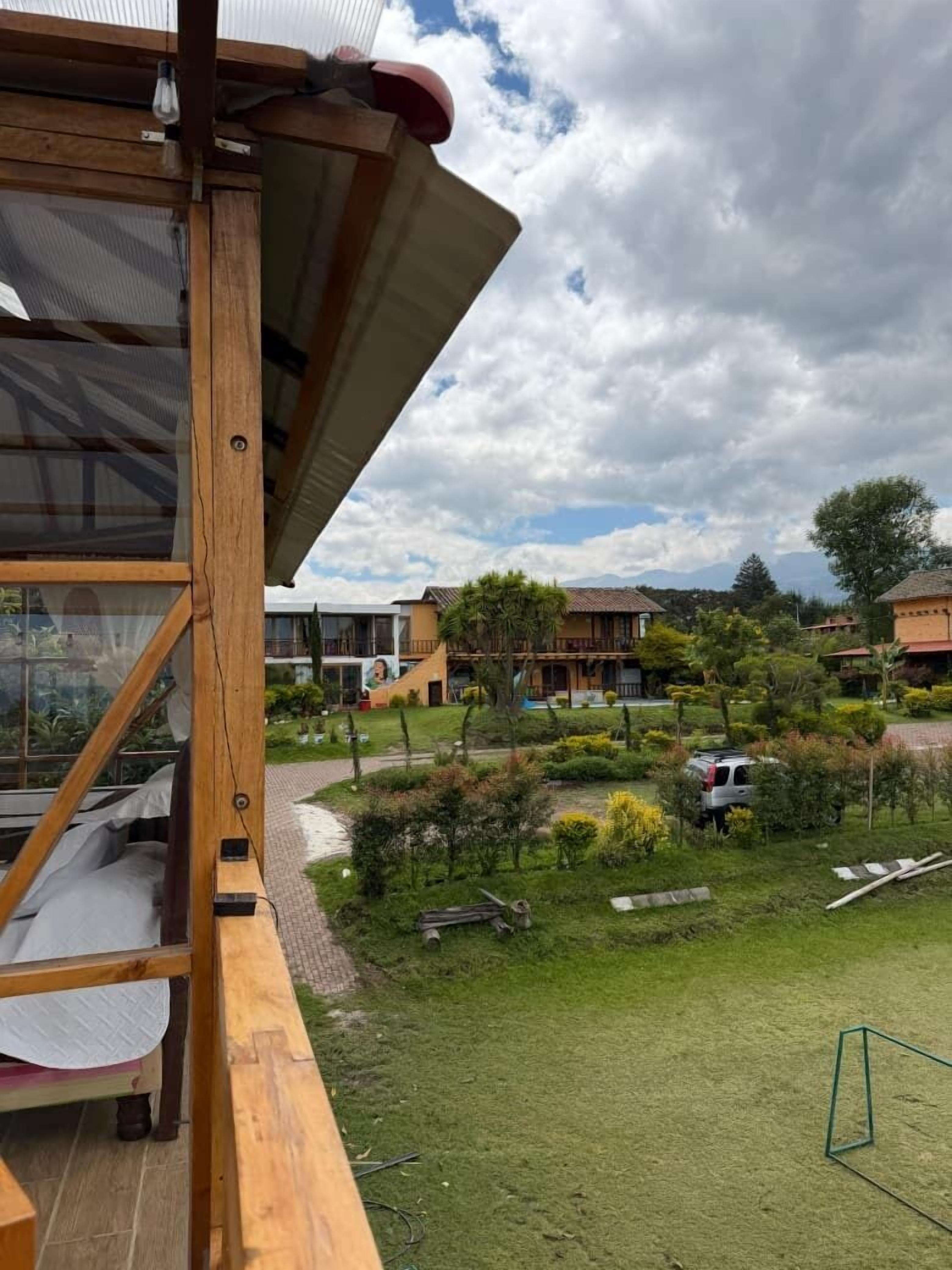Una terraza de madera con barandilla de cristal que da a un jardín ajardinado con un camino y vehículos estacionados.