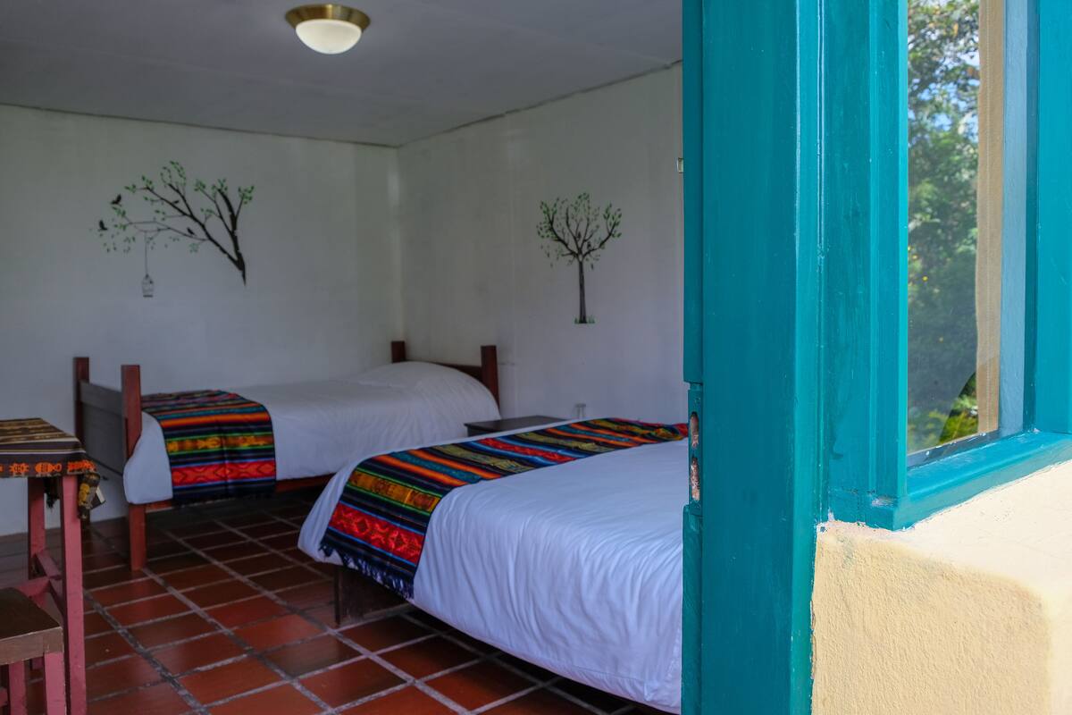 A room with two beds, a small table, and a window with a view of trees.