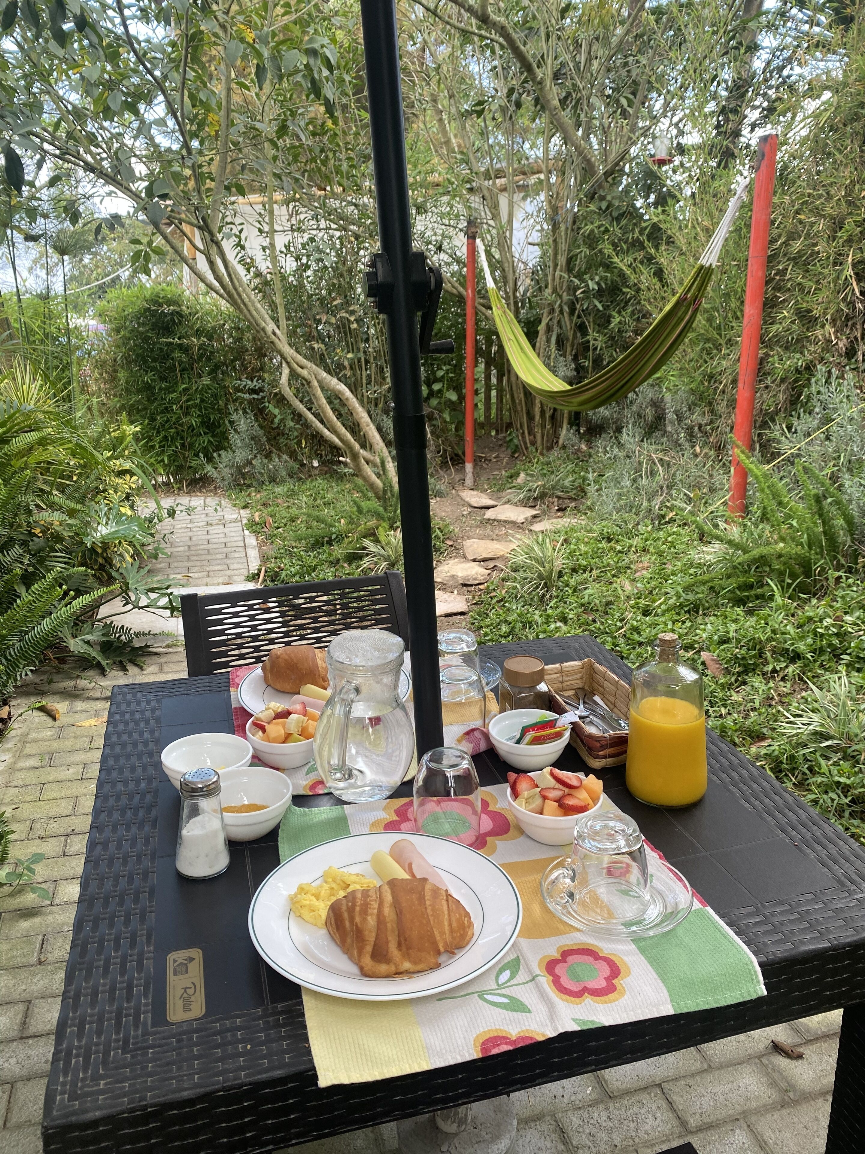 An outdoor dining table set with breakfast items, surrounded by greenery and a hammock.