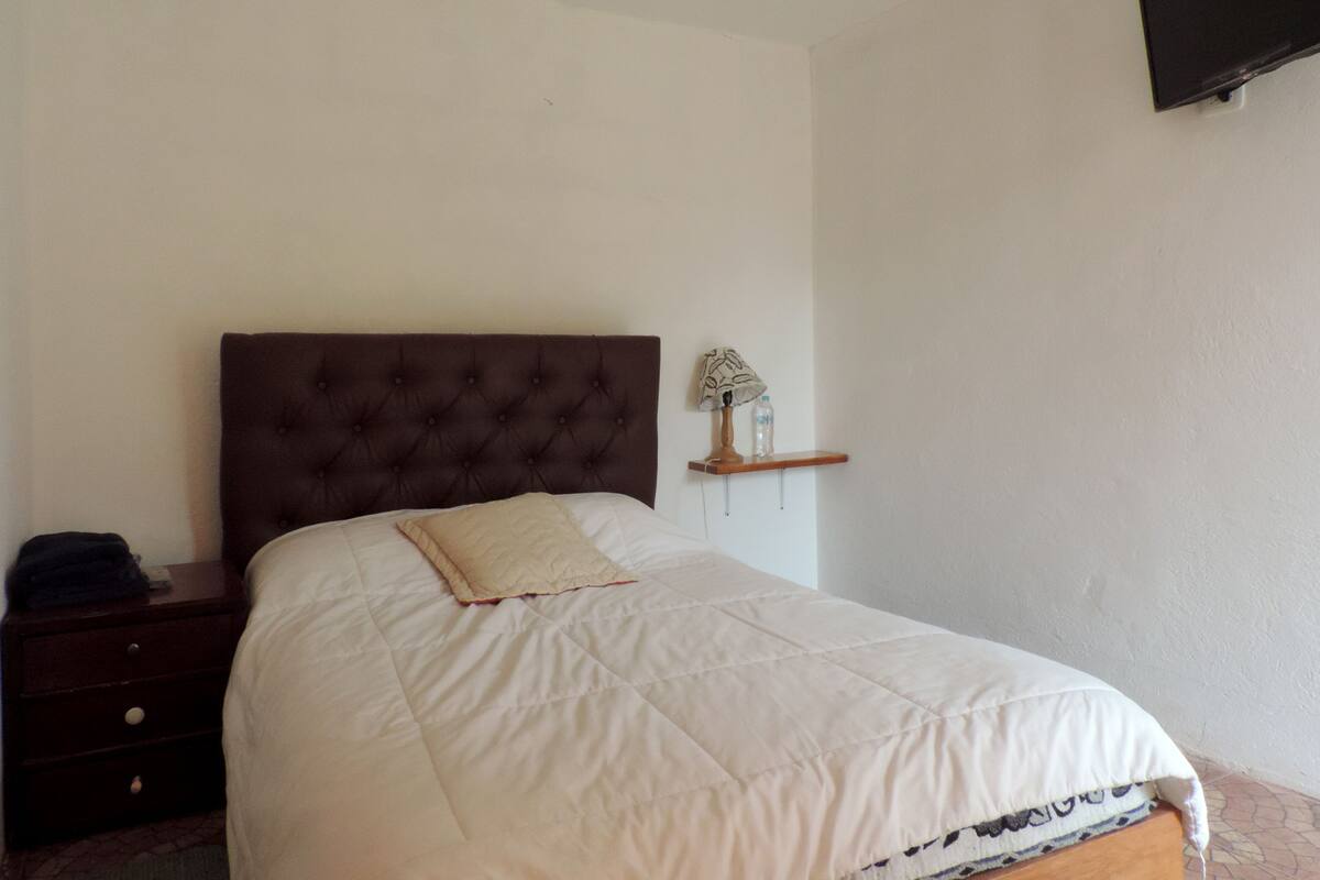 A bedroom with a large bed, a nightstand, a wall-mounted TV, and a small shelf with a lamp and a bottle.