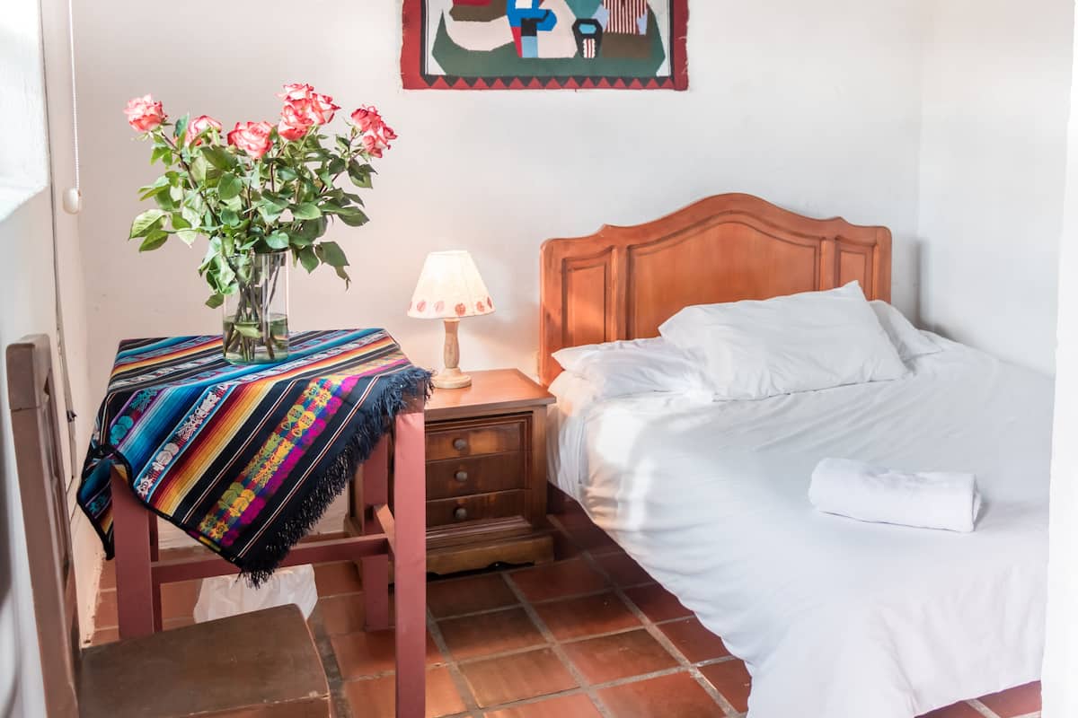A hotel room with a bed, a nightstand with a lamp, a chair with a colorful blanket, a vase with flowers, and a wall hanging.