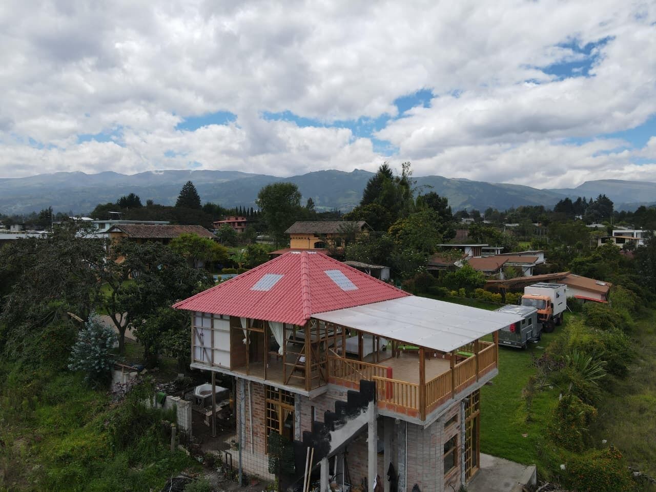 Una casa de dos pisos con techo rojo, rodeada de vegetación y montañas al fondo.