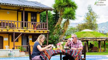A dining table with a colorful striped tablecloth, a pool, and a yellow building with a balcony.