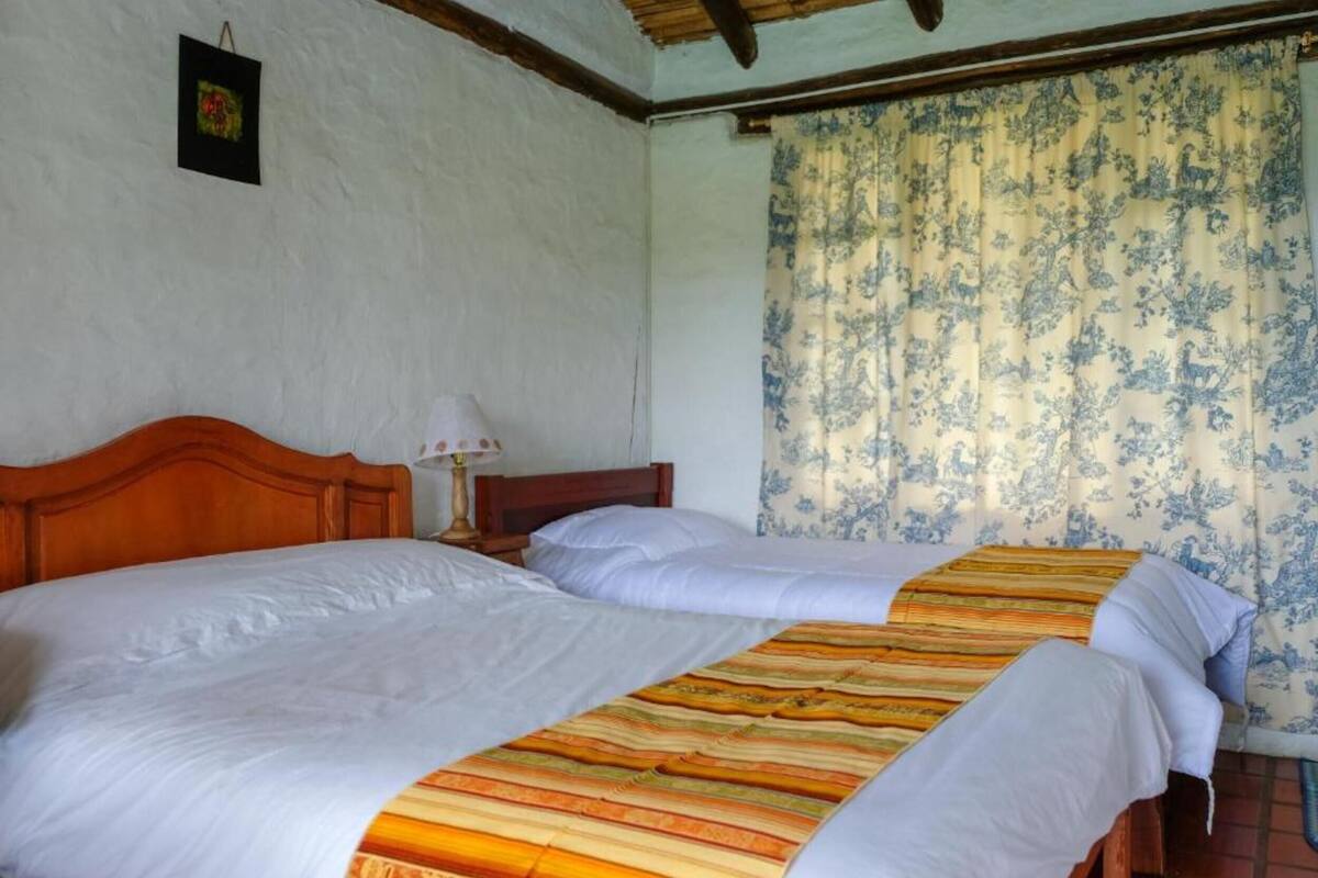A room with two beds, a wooden headboard, a nightstand with a lamp, and a floral curtain.