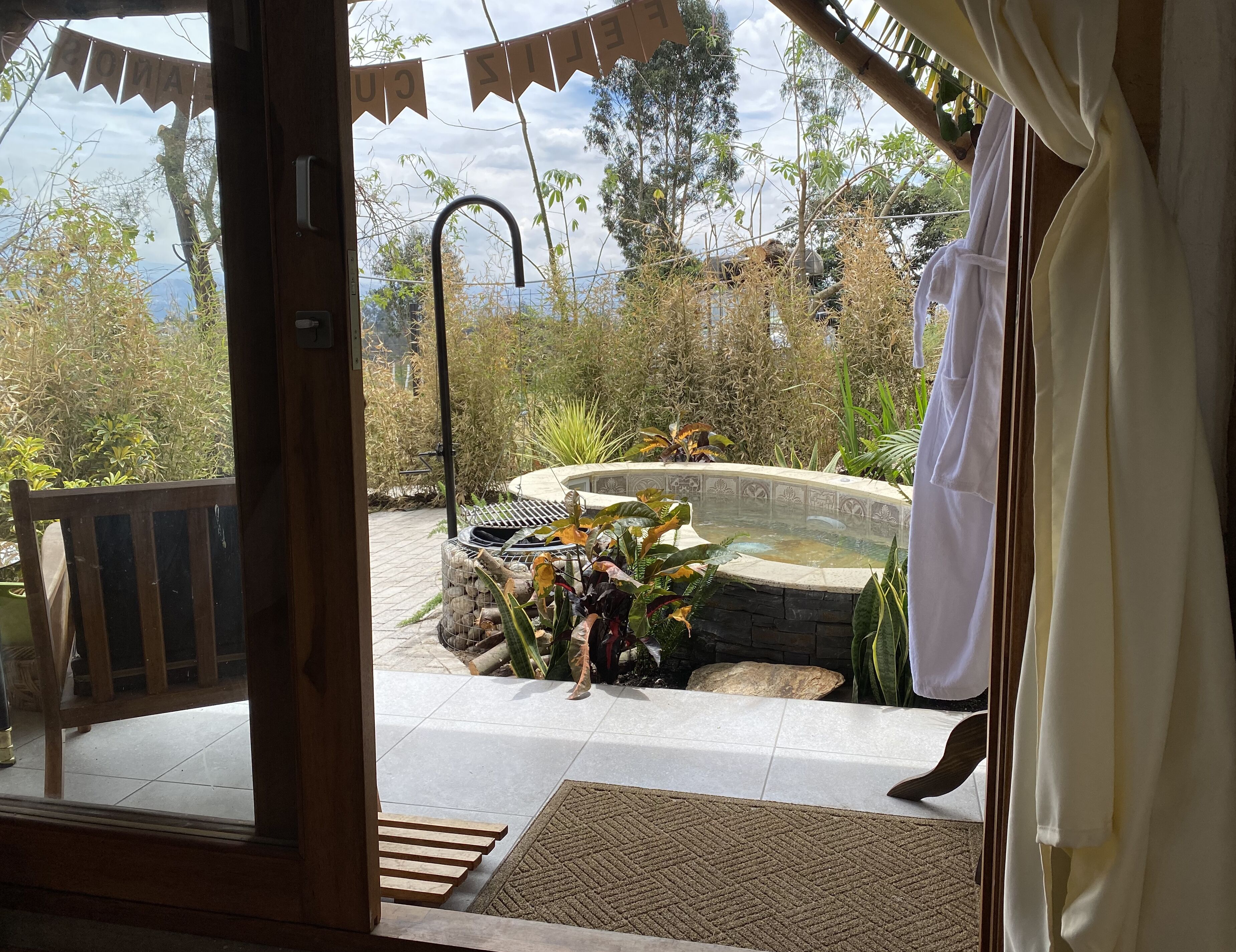 A view from inside a room to an outdoor garden area with a hot tub, surrounded by plants and a wooden deck.