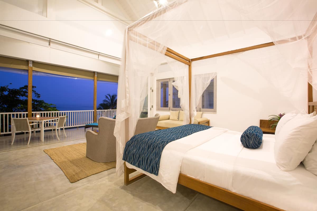 A bedroom with a four-poster bed, a balcony with a table and chairs, and a view of the ocean at dusk.