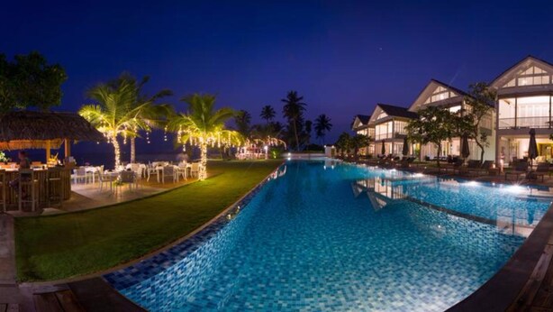A swimming pool with a wooden deck, a thatched-roof structure, and a building with large windows. - Sri Sharavi Beach Villas & Spa (Thalaramba)