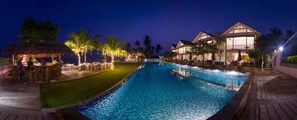 A swimming pool with a wooden deck, a thatched-roof structure, and a building with large windows. - Sri Sharavi Beach Villas & Spa (Thalaramba)