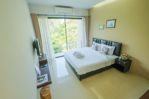 A hotel room with a large bed, a nightstand with a clock, and a view of greenery through the window.
