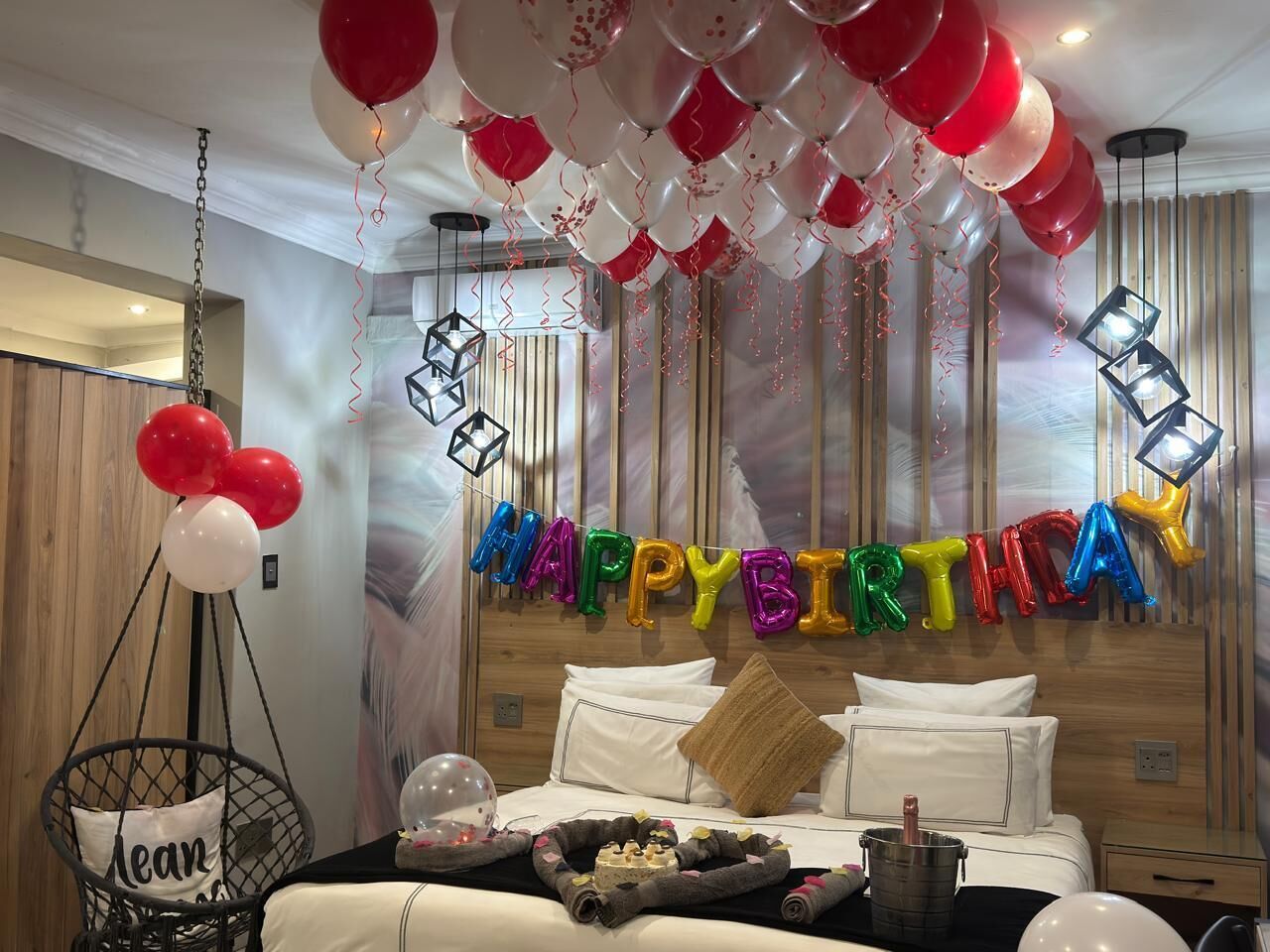 A room with a bed, a hanging chair, and a wall decorated with balloons and a 'HAPPY BIRTHDAY' sign.