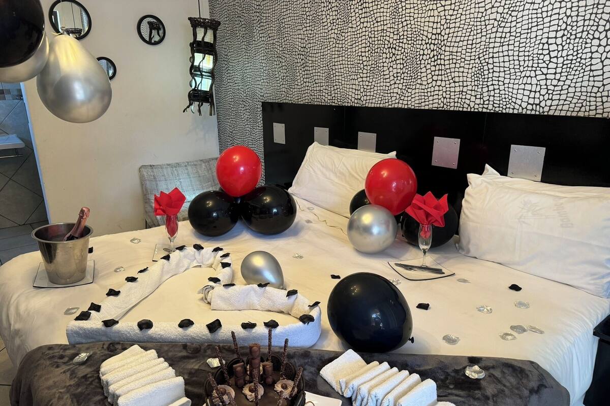 Een bed met een hoofdbord in zwart-wit patroon, ballonnen, een champagnefles en een chocoladetaart.