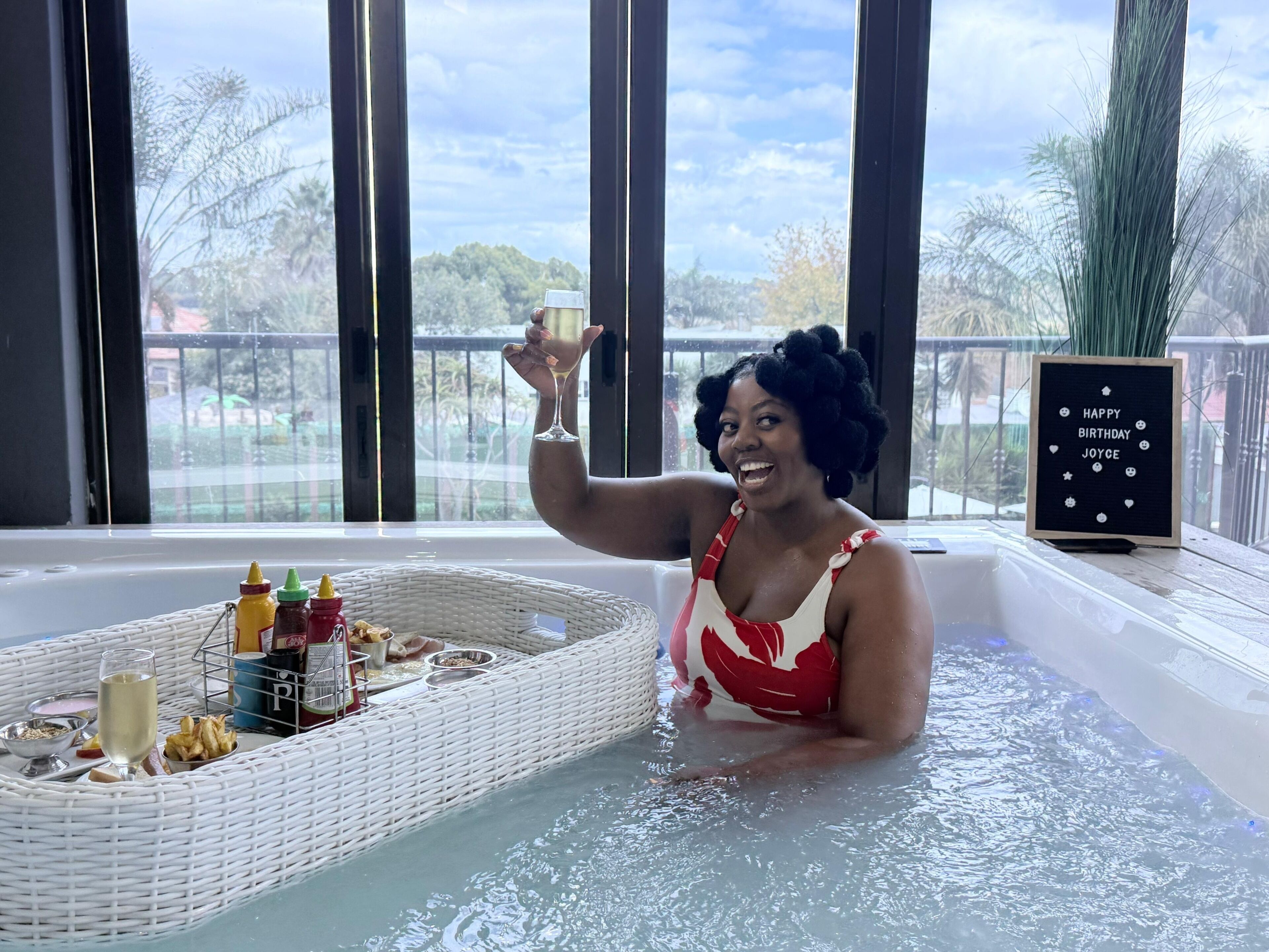 Una persona en un jacuzzi con una bandeja de comida y bebidas, un cartel con un mensaje de cumpleaños y vistas a árboles y una valla.
