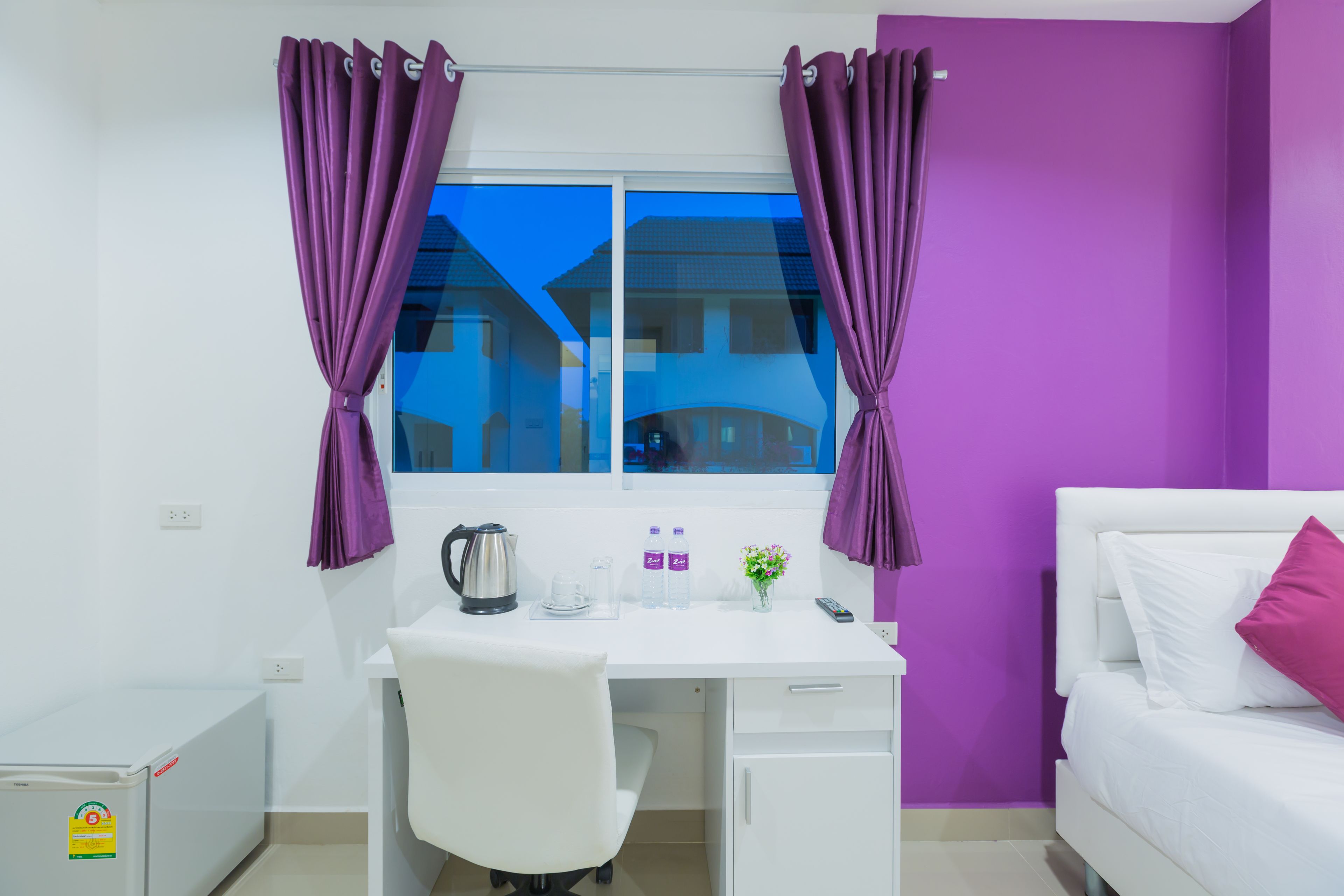 Une pièce avec un bureau blanc, des rideaux violets, un lit avec des oreillers blancs et roses, et une fenêtre donnant sur des immeubles.