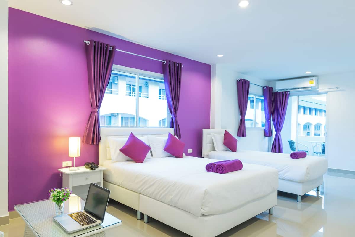 A hotel room with two beds, purple curtains, a laptop on a glass table, and a view of buildings outside.