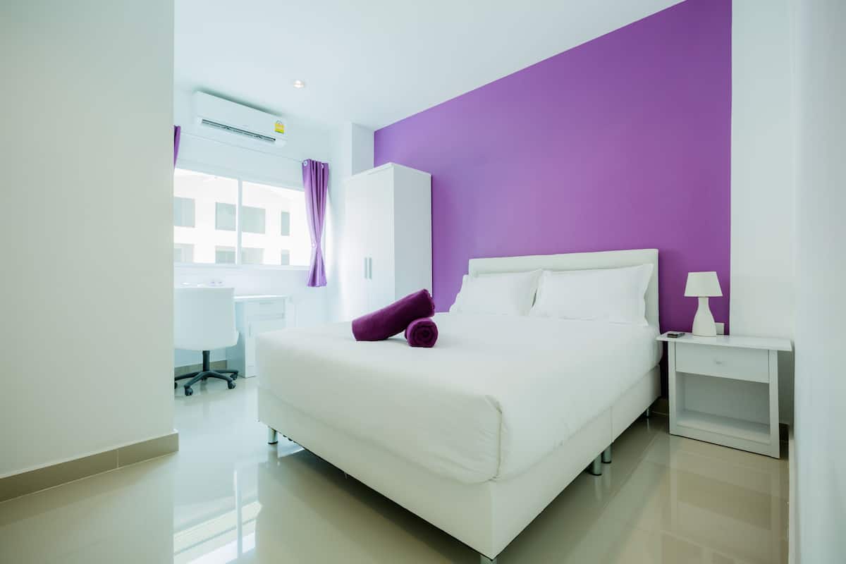 A modern hotel room with a large bed, white walls, and a purple accent wall.