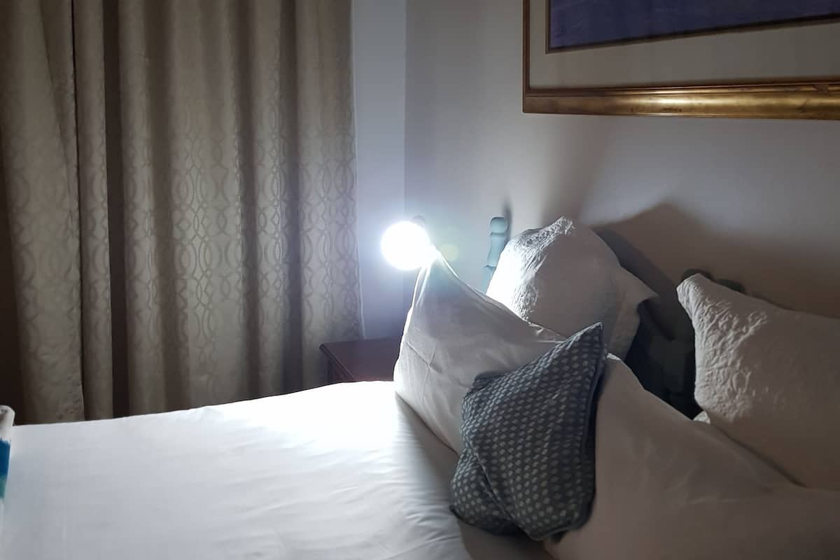 Una habitación de hotel con cama, almohadas, una mesita de noche, una lámpara y un cuadro en la pared.