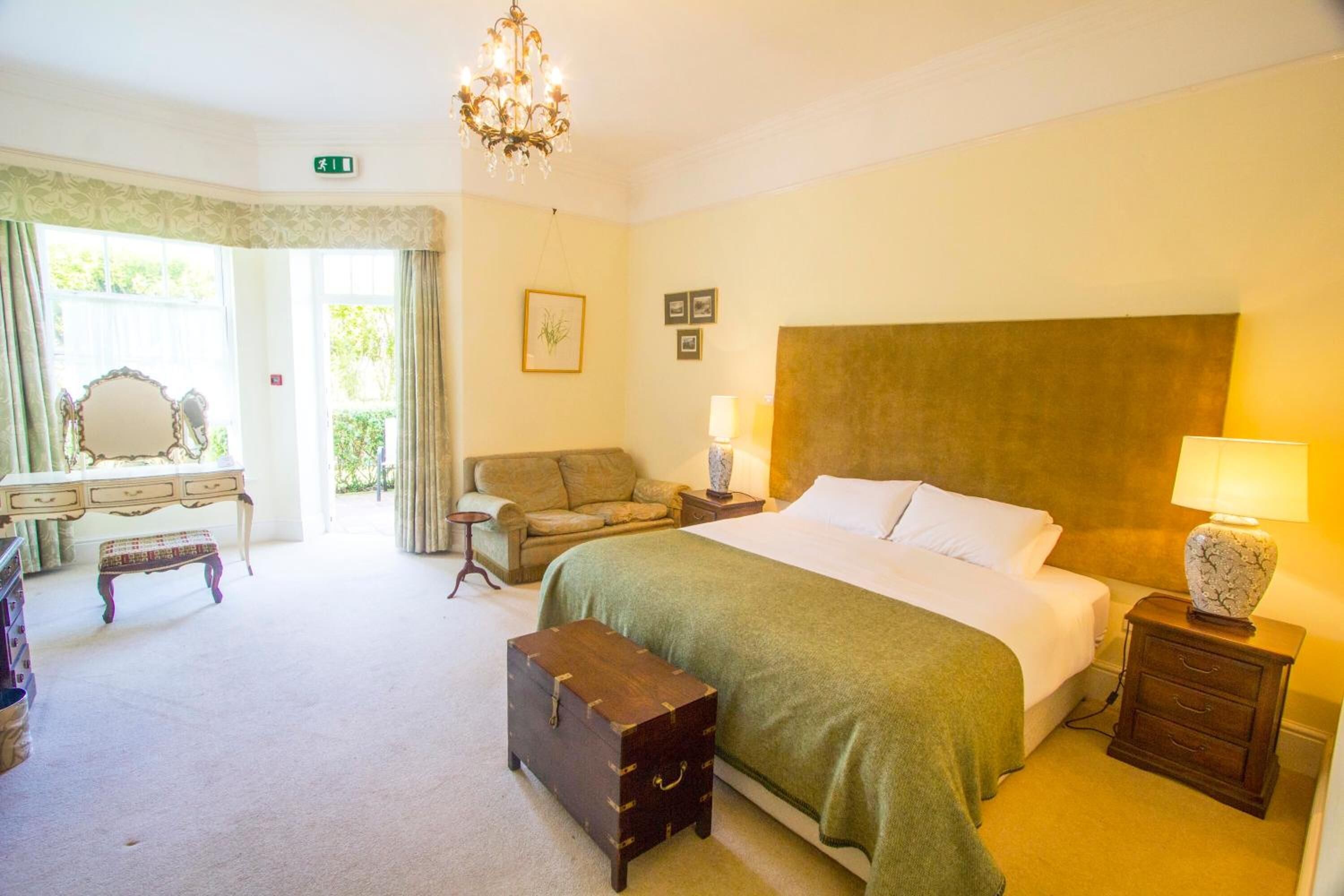 A hotel room with a large bed, bedside tables, a bench, a dresser, and a chandelier.