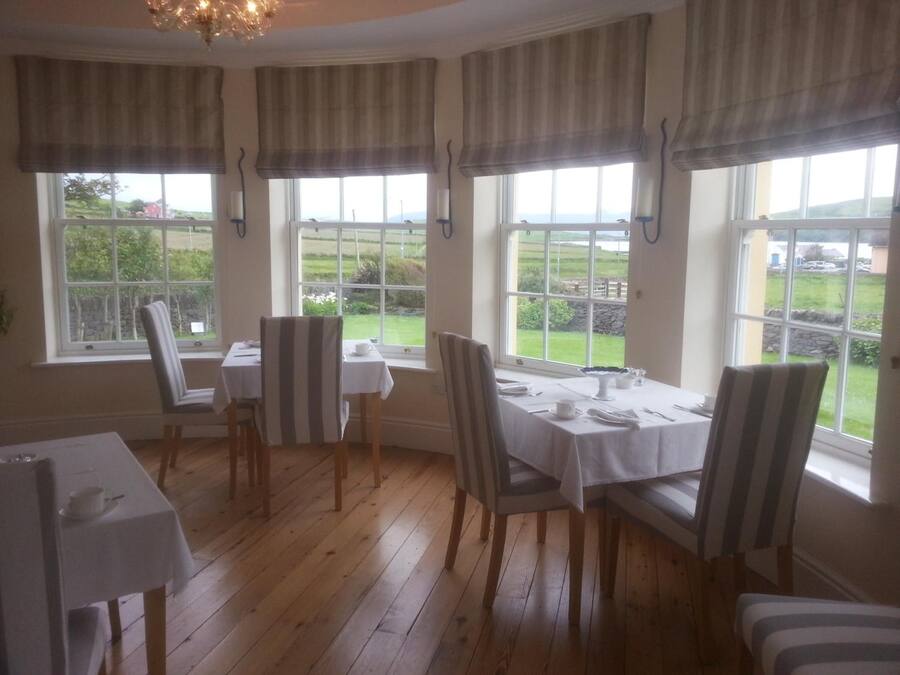 A dining area with wooden flooring, striped chairs, and tables set for four. Large windows with curtains offer a view of the outdoors.
