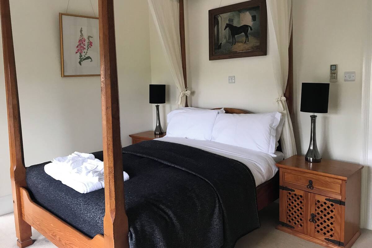A four-poster bed with white linens, two bedside tables with lamps, a wooden nightstand, and a painting of a horse on the wall.