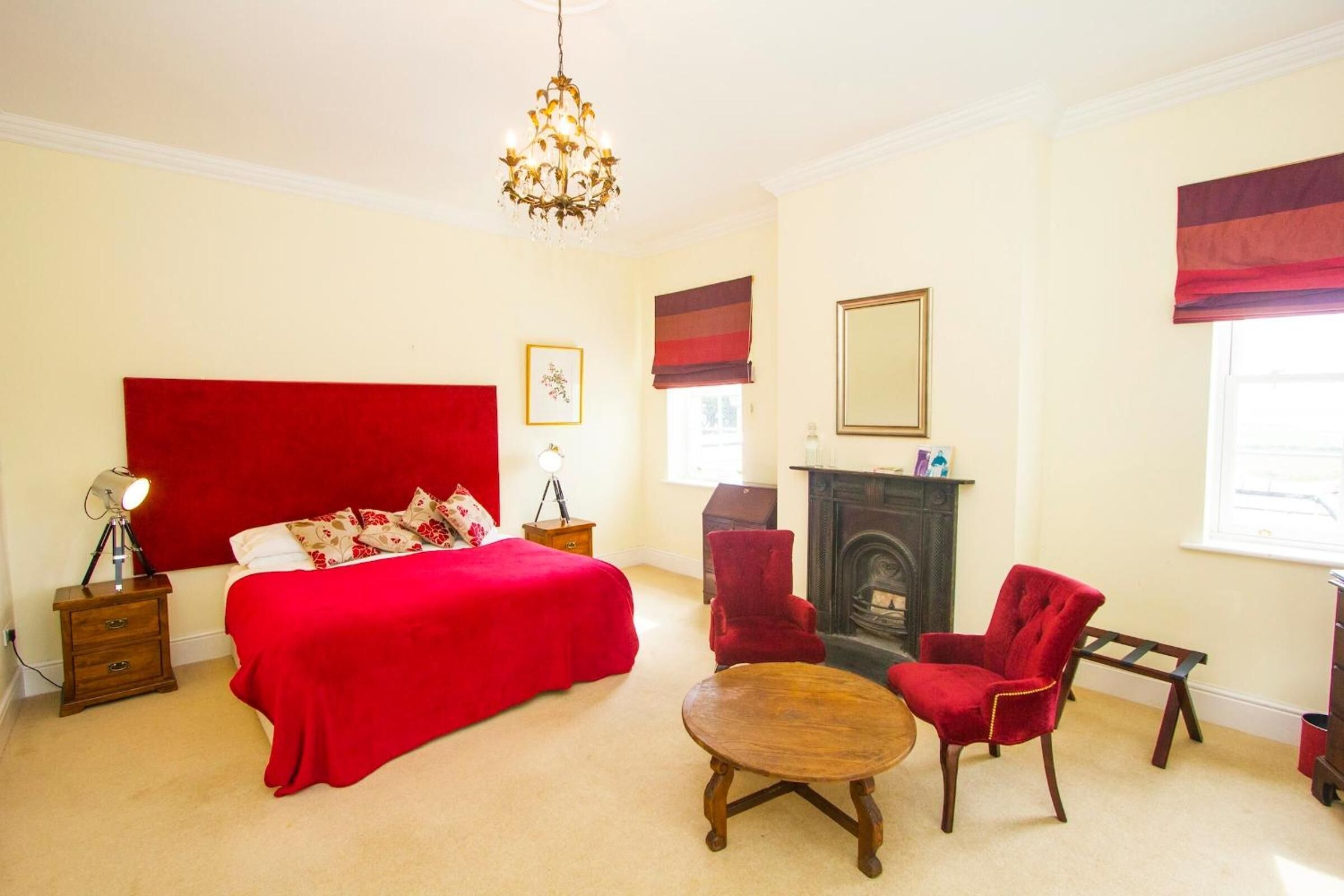 A bedroom with a large bed, a fireplace, a chandelier, and red upholstered chairs.