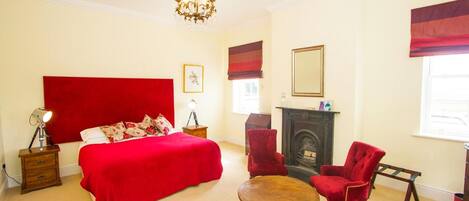 A bedroom with a large bed, a fireplace, a chandelier, and red upholstered chairs.