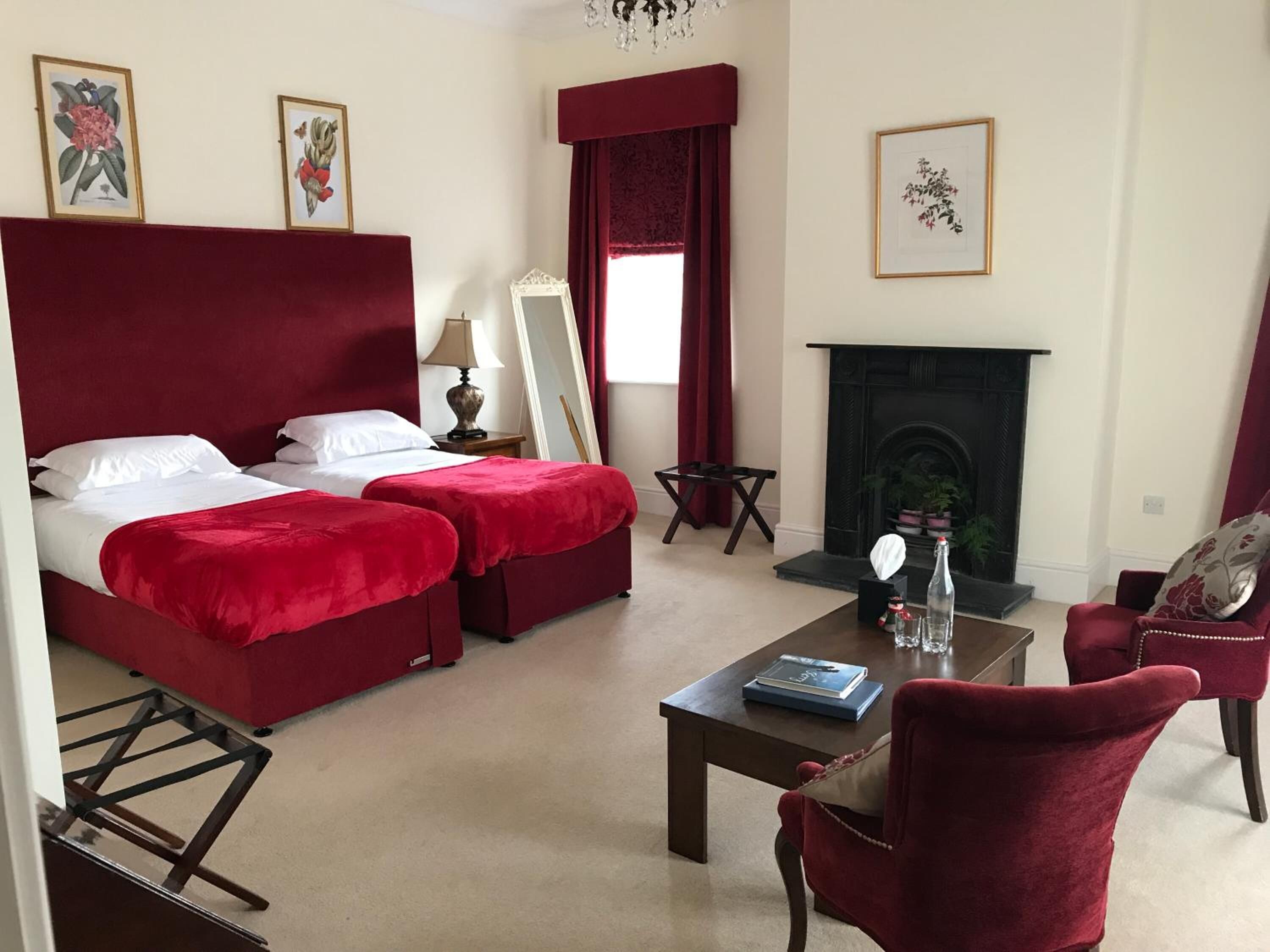 Habitación de hotel con una cama grande, dos sillones tapizados en rojo, una mesita con un libro, chimenea e impresiones botánicas enmarcadas en las paredes.
