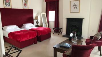 Habitación de hotel con una cama grande, dos sillones tapizados en rojo, una mesita con un libro, chimenea e impresiones botánicas enmarcadas en las paredes.
