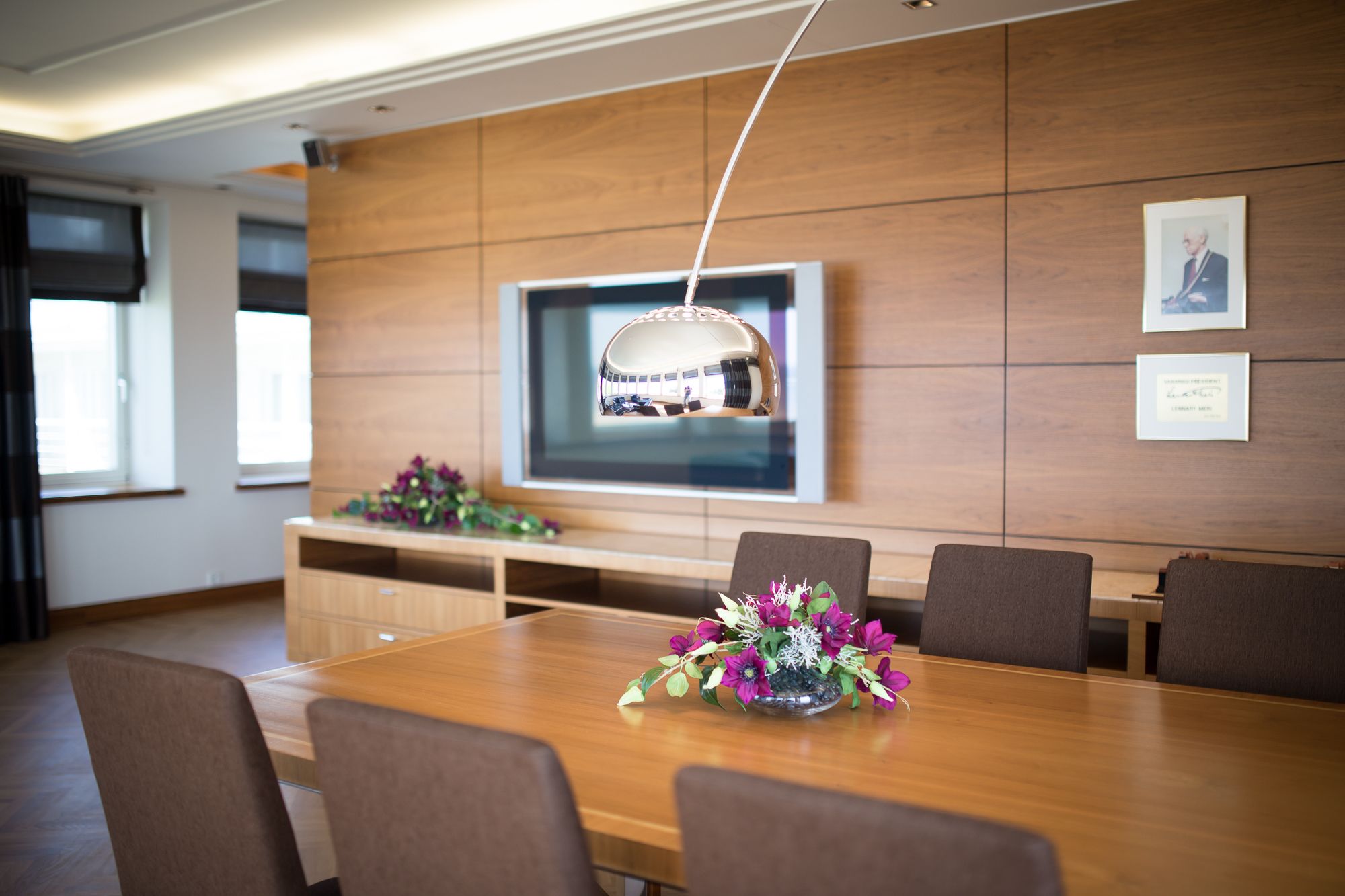 Un salón de conferencias con una larga mesa, seis sillas, un televisor de pantalla plana grande montado en la pared y un centro de mesa floral.