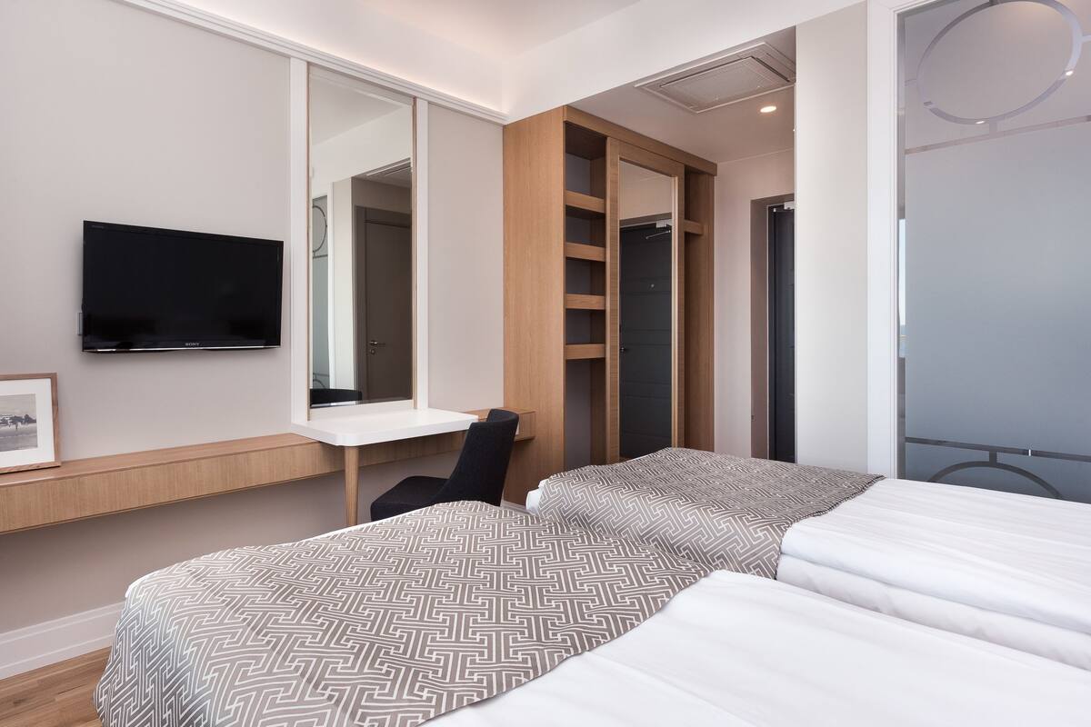 A modern hotel room with a flat-screen TV, a desk, a chair, a bed with patterned bedding, and a large mirror.