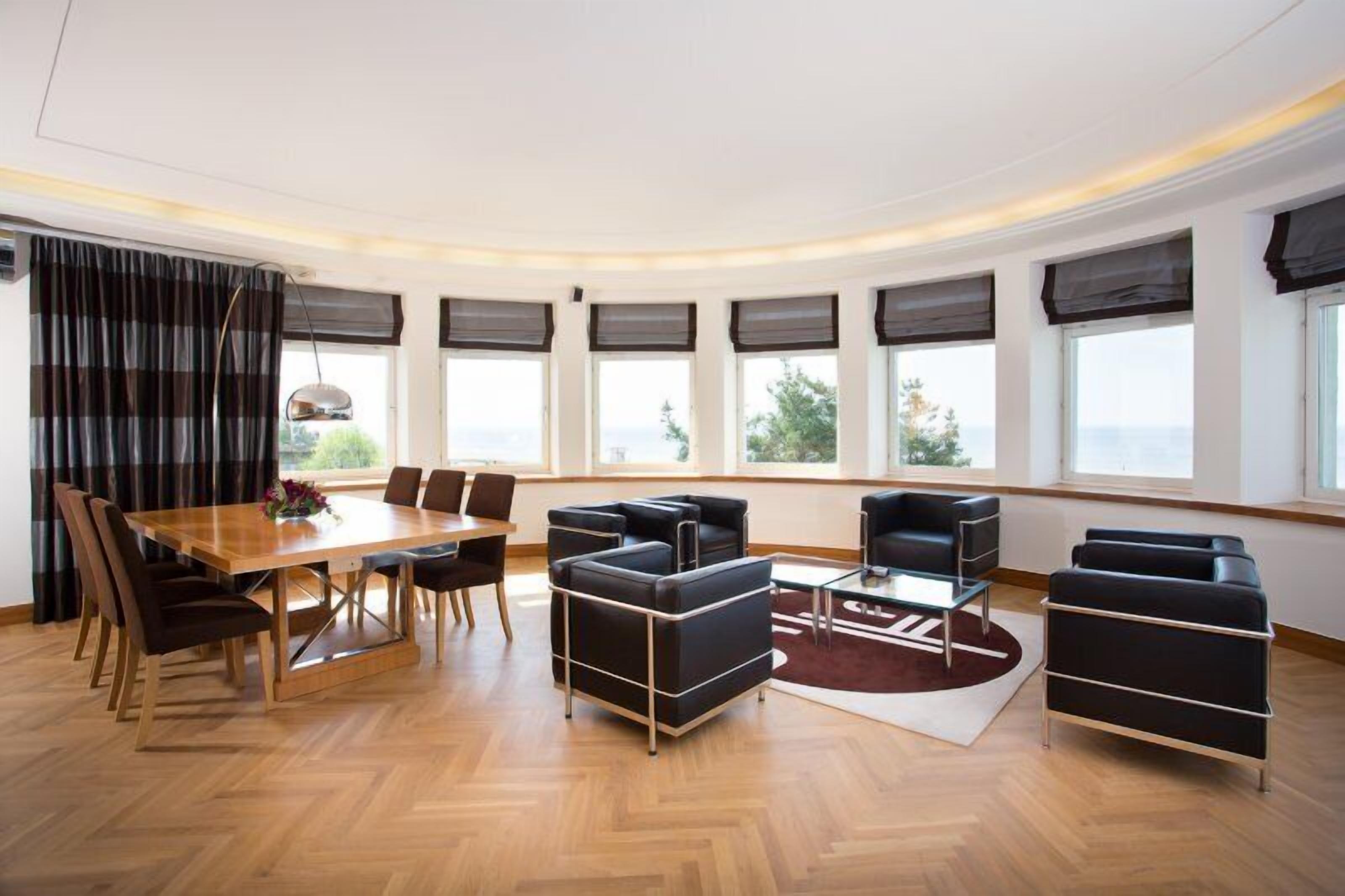 Ein modernes Wohnzimmer mit einem Holzesstisch, schwarzen Sofas und großen Fenstern mit Vorhängen.