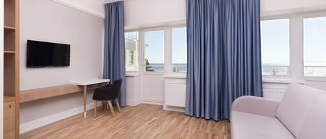 A modern room with a wooden floor, a white desk with a chair, a flat-screen TV, a sofa, and large windows with blue curtains.
