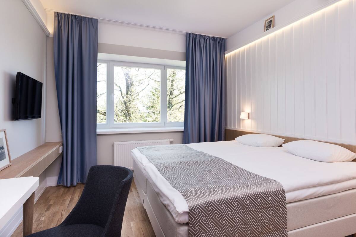 A hotel room with a bed, bedside table, chair, and a window with curtains.