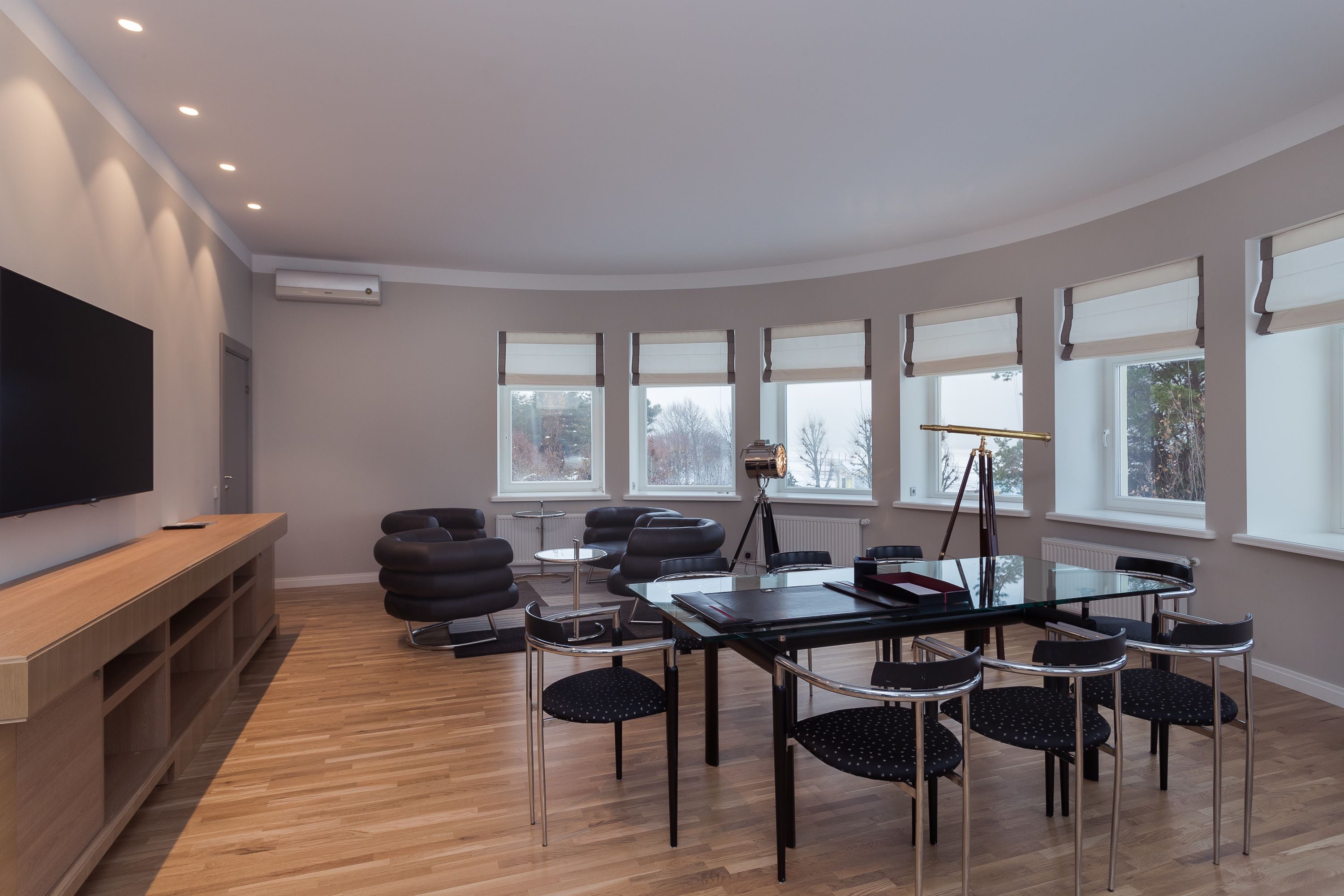 Una habitación moderna con piso de madera, una mesa de vidrio y sillas negras. Hay un televisor de pantalla plana en un estante de madera, y una ventana con vistas a árboles.