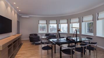 Una habitación moderna con piso de madera, una mesa de vidrio y sillas negras. Hay un televisor de pantalla plana en un estante de madera, y una ventana con vistas a árboles.