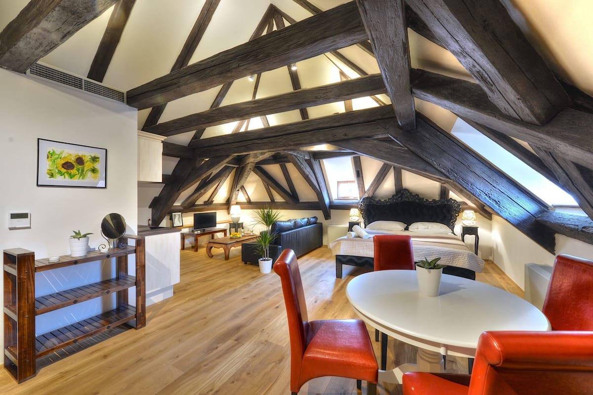 An attic room with exposed wooden beams, a round dining table, red chairs, a sofa, a bed, and a small table with a lamp.